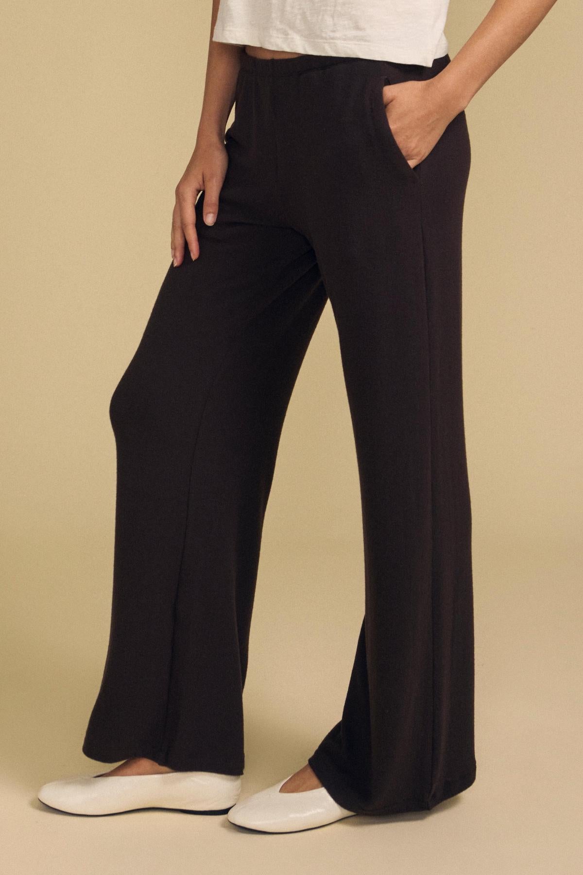 A person wearing the MACKENZIE LOUNGE PANT by Velvet by Graham & Spencer in black, styled with a white shirt and white flats, stands with one hand in their pocket against a beige background.