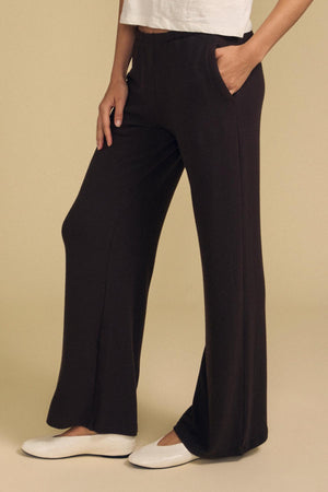A person wearing the MACKENZIE LOUNGE PANT by Velvet by Graham & Spencer in black, styled with a white shirt and white flats, stands with one hand in their pocket against a beige background.