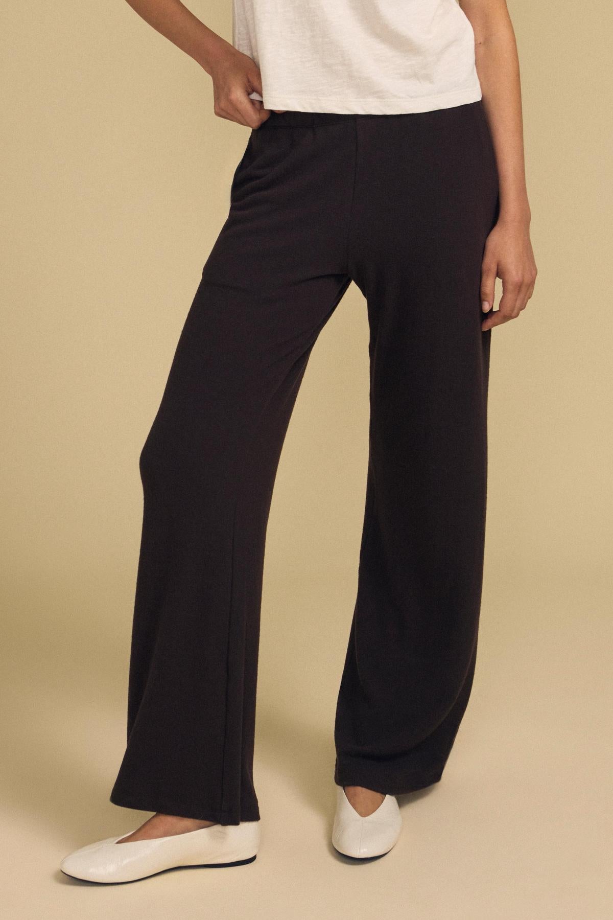 Model wears the MACKENZIE LOUNGE PANT by Velvet by Graham & Spencer in black, paired with a white short-sleeve top and white flat shoes, standing against a plain beige background.