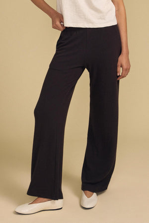 Model wears the MACKENZIE LOUNGE PANT by Velvet by Graham & Spencer in black, paired with a white short-sleeve top and white flat shoes, standing against a plain beige background.