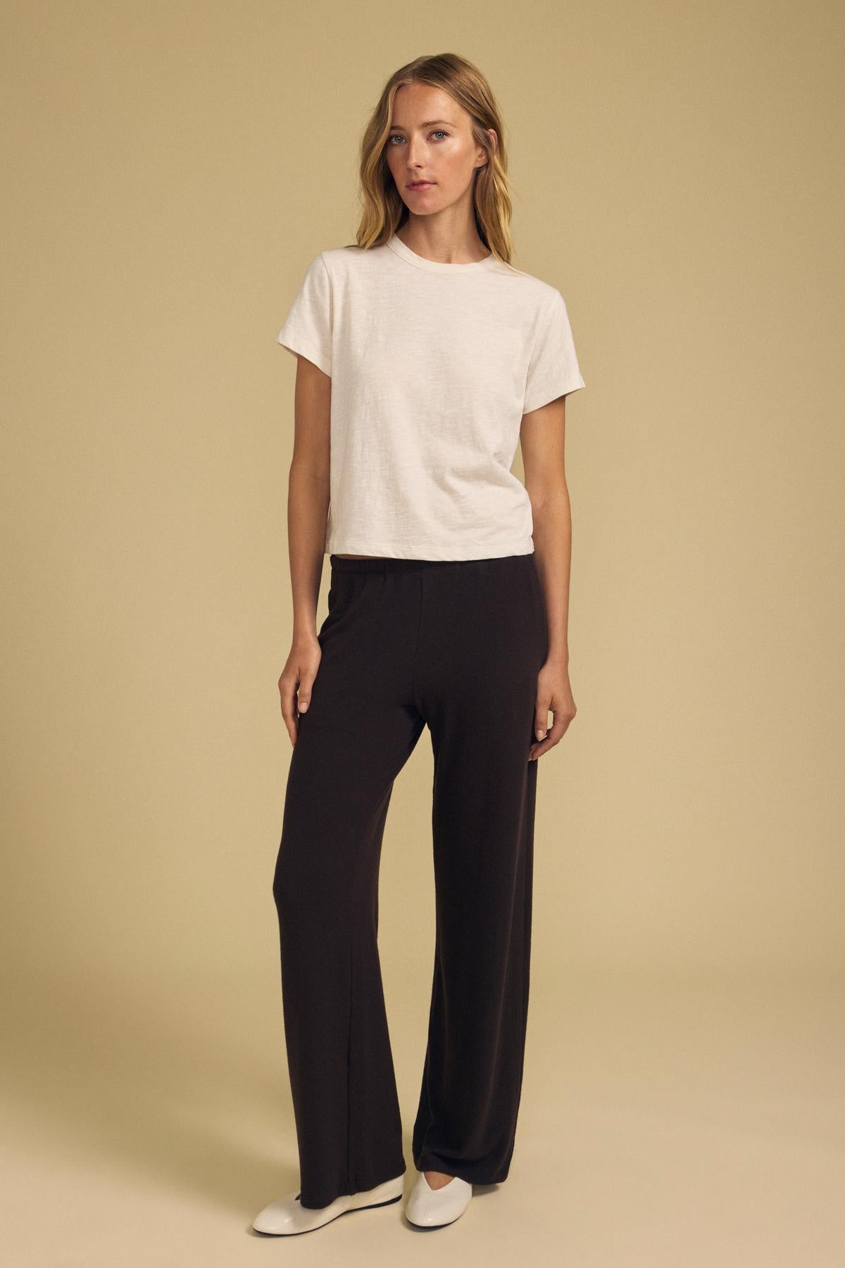 A woman stands against a beige background, wearing a white short-sleeve t-shirt, the MACKENZIE LOUNGE PANT in black by Velvet by Graham & Spencer, and white shoes.-42092007555265