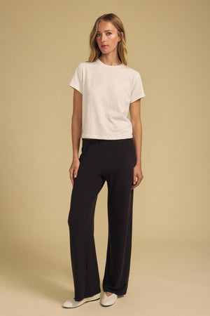 A woman stands against a beige background, wearing a white short-sleeve t-shirt, the MACKENZIE LOUNGE PANT in black by Velvet by Graham & Spencer, and white shoes.