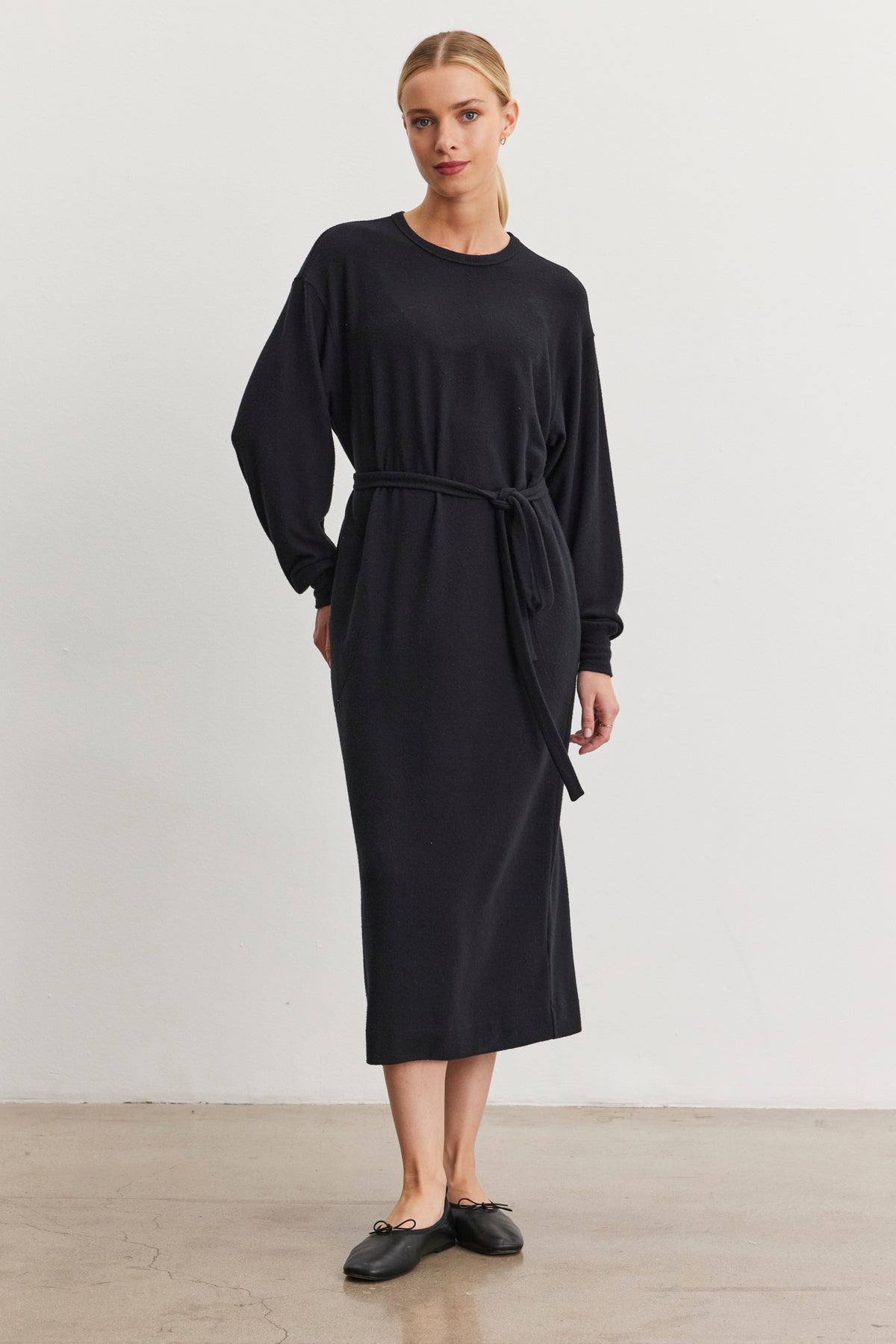   In a minimalist setting, a person stands effortlessly stylish in the AIDA DRESS by Velvet by Graham & Spencer, a long and elegant black triblend knit dress featuring a detachable belt. Their ensemble is completed with sleek black shoes, and their light-colored hair is neatly tied back. 