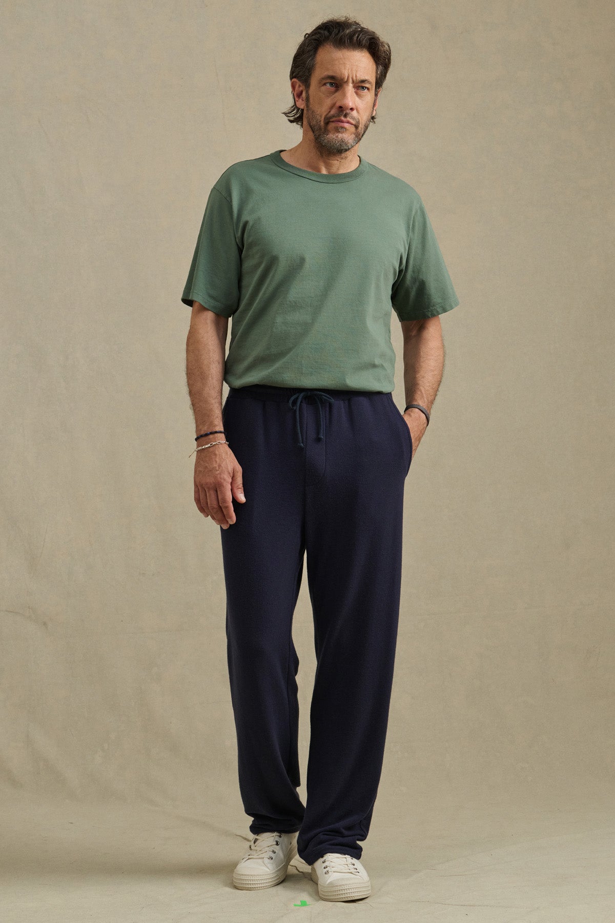 A man stands facing forward wearing the EASTON SWEATPANT by Velvet by Graham & Spencer in dark blue with a green t-shirt and white sneakers, set against a plain beige background.-41009006149825