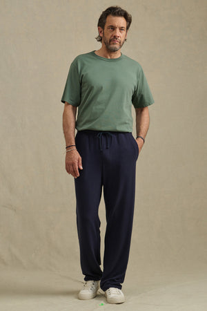 A man stands facing forward wearing the EASTON SWEATPANT by Velvet by Graham & Spencer in dark blue with a green t-shirt and white sneakers, set against a plain beige background.