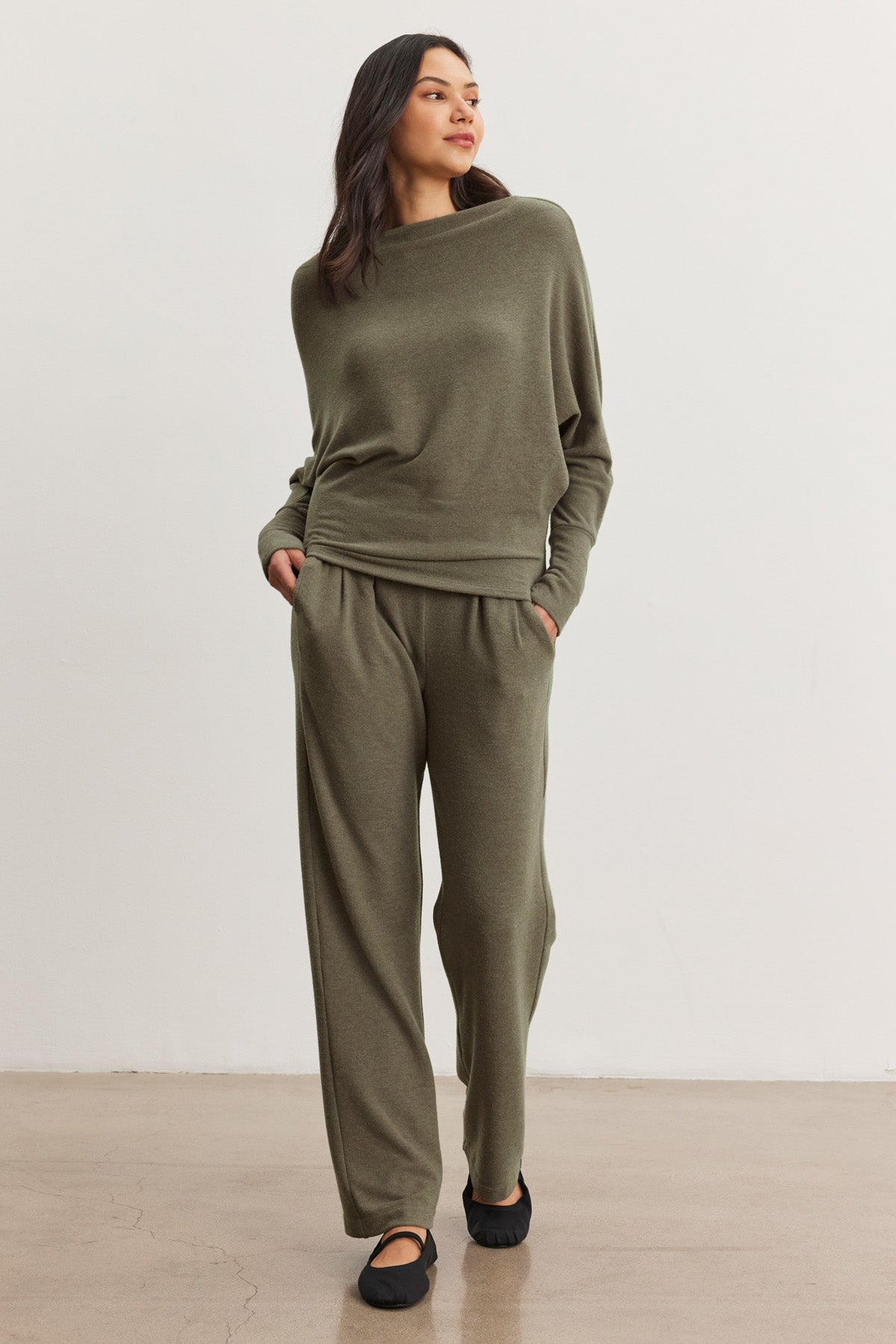 A person stands indoors, hands in pockets, wearing the Velvet by Graham & Spencer ELIYA SWEATPANT in olive green with a matching long-sleeve top, looking to the side.