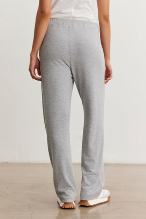 A rear view showcases someone wearing the ELIYA PANT from Velvet by Graham & Spencer. These gray sweatpants feature a straight leg design and offer a comfortable fit, complemented by a white shirt, as they stand on a polished floor.