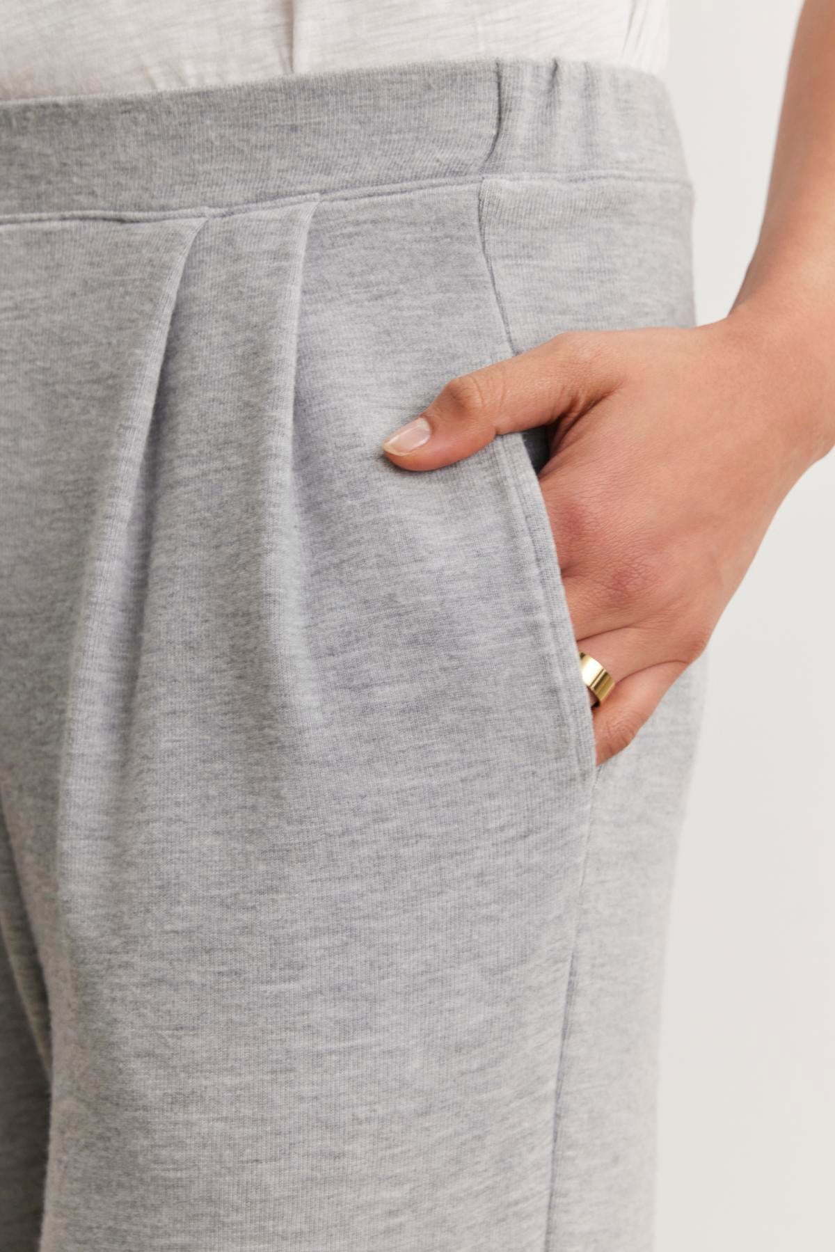   Close-up of an individual wearing the ELIYA PANT by Velvet by Graham & Spencer, featuring a gray triblend knit with a straight leg design and their hand casually resting in the pocket. A ring shines on their finger, enhancing the pant's comfortable style. 