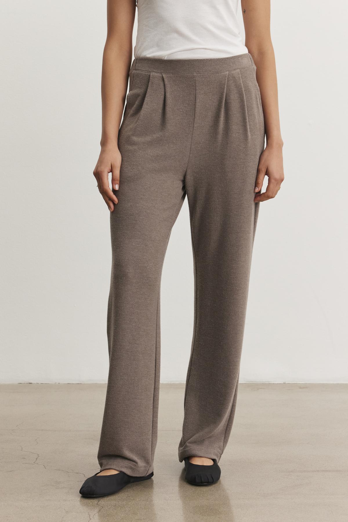 A person wearing the ELIYA PANT by Velvet by Graham & Spencer, which features a taupe color and a comfortable wide-leg fit, along with a white top, stands on a polished concrete floor.-38433293697217