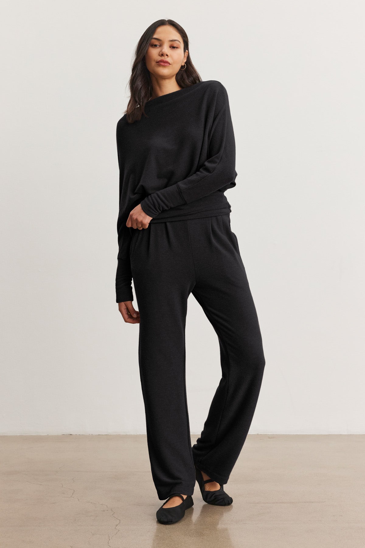 A woman stands against a plain background wearing the Velvet by Graham & Spencer SELMY TOP, a loose-fitting black triblend knit long-sleeve top with dolman sleeves, paired with matching black wide-leg pants and black flats.-40461709181121