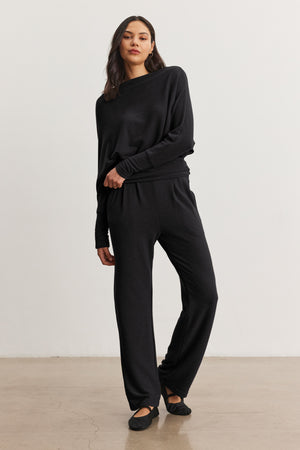 A woman stands against a plain background wearing the Velvet by Graham & Spencer SELMY TOP, a loose-fitting black triblend knit long-sleeve top with dolman sleeves, paired with matching black wide-leg pants and black flats.
