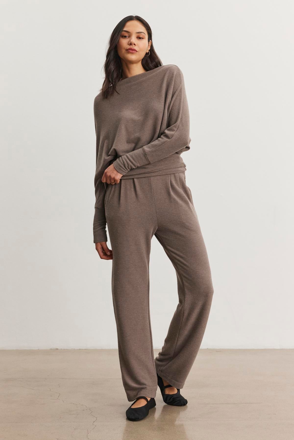   A woman stands on a plain background wearing the SELMY TOP by Velvet by Graham & Spencer, a soft triblend knit lounge outfit featuring dolman sleeves and a long-sleeved taupe top with matching pants and black shoes. 