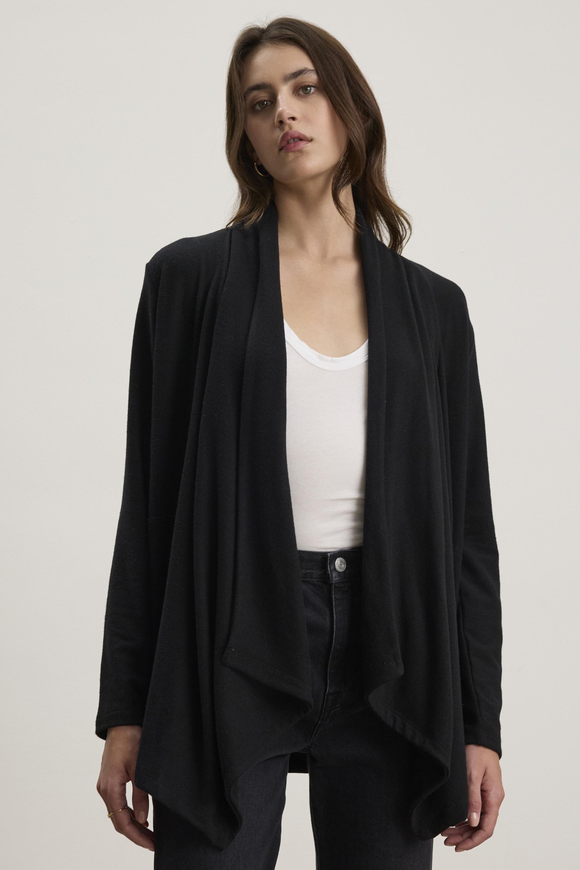   A person is wearing the Velvet by Graham & Spencer LISA CARDIGAN, a black triblend knit cardigan with an open front, over a white top and black pants, standing against a plain background. 