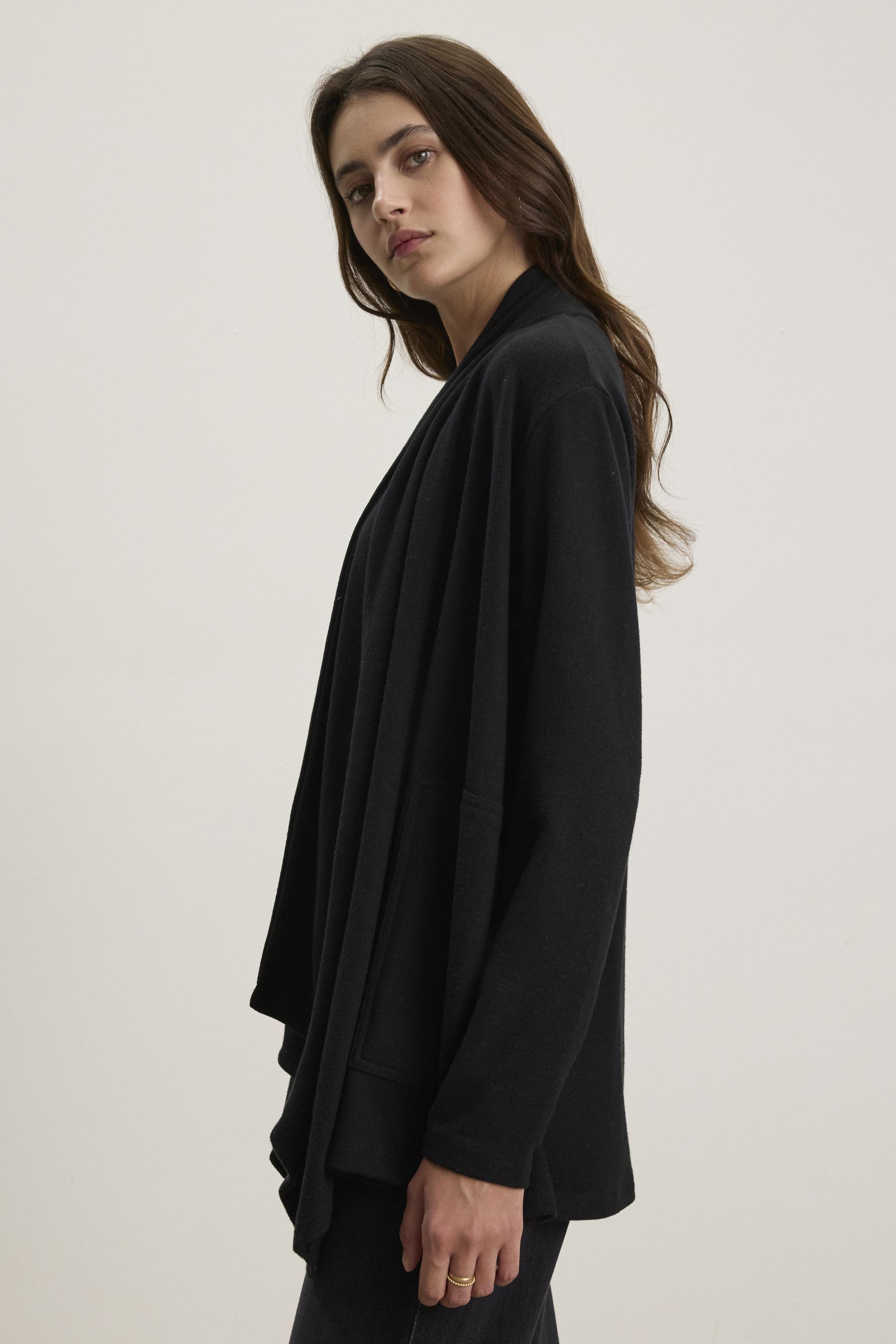   A person with long hair is wearing the LISA CARDIGAN by Velvet by Graham & Spencer, a black triblend knit cardigan with an open front and side patch pockets, standing against a plain background. 