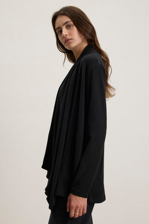 A person with long hair is wearing the LISA CARDIGAN by Velvet by Graham & Spencer, a black triblend knit cardigan with an open front and side patch pockets, standing against a plain background.