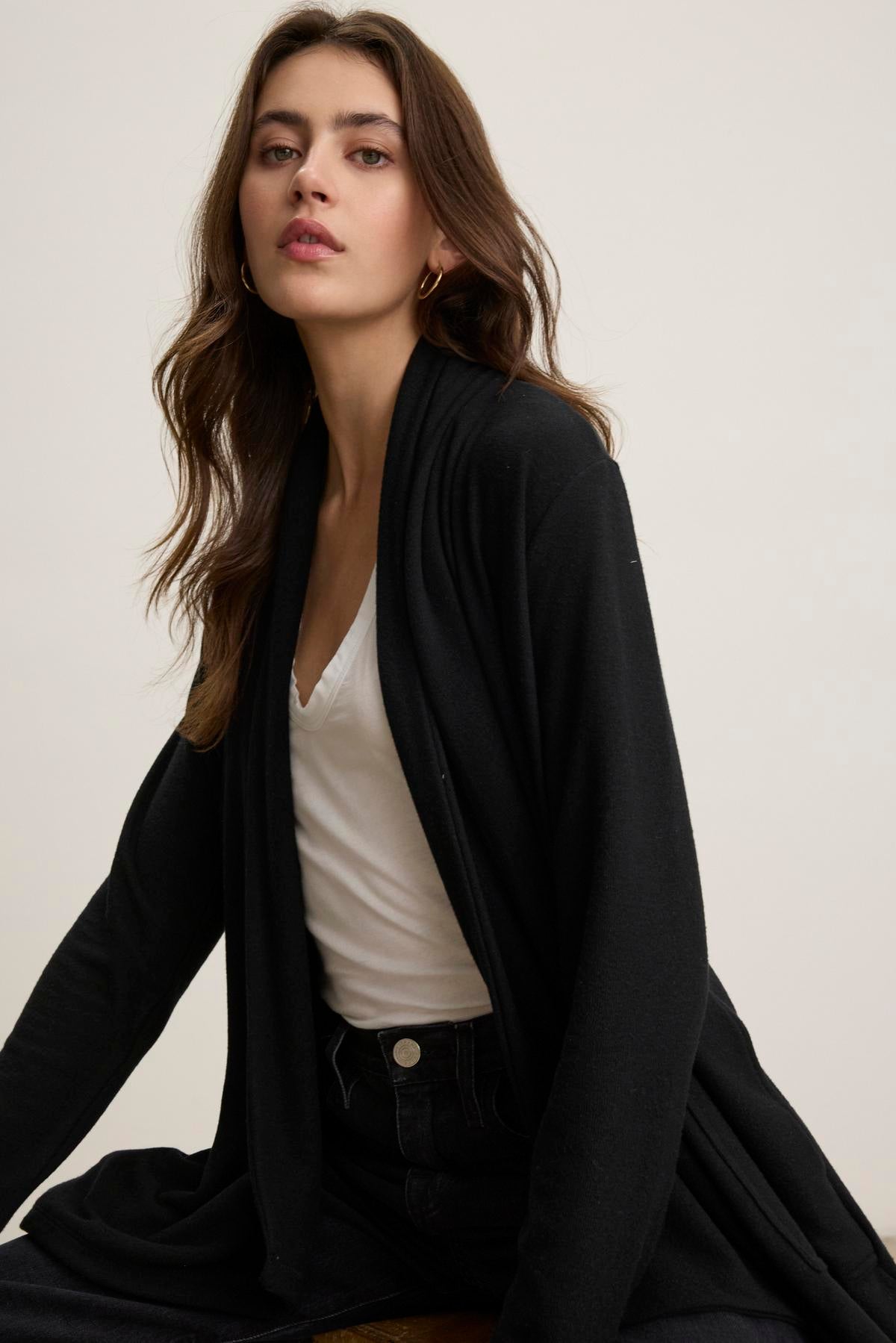   A woman wearing a white V-neck top along with a LISA CARDIGAN by Velvet by Graham & Spencer sits with a neutral expression, gazing at the camera against a simple backdrop. 