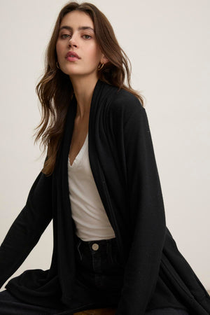 A woman wearing a white V-neck top along with a LISA CARDIGAN by Velvet by Graham & Spencer sits with a neutral expression, gazing at the camera against a simple backdrop.