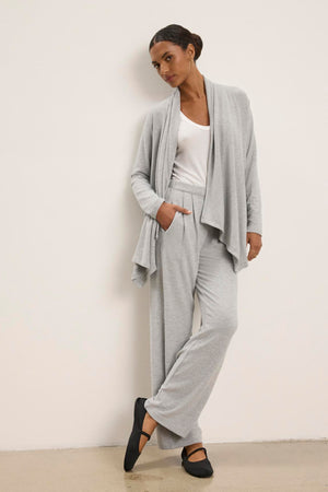 A person leans against a wall wearing Velvet by Graham & Spencer's light gray triblend knit cardigan, ELIYA PANT in a matching wide-leg style, a white top, and black shoes for a comfortable fit.