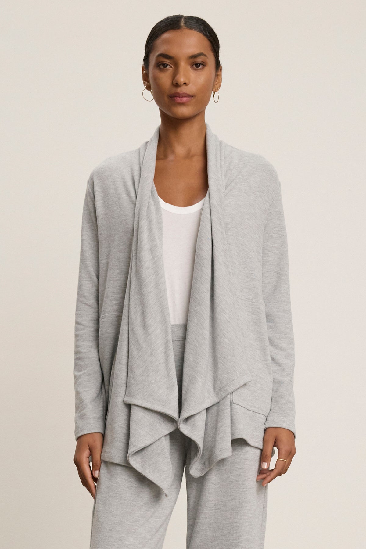   A person wearing a grey LISA CARDIGAN by Velvet by Graham & Spencer, crafted from luxurious triblend knit fabric with side patch pockets, is styled over a white shirt and matching grey pants, standing against a neutral background. 