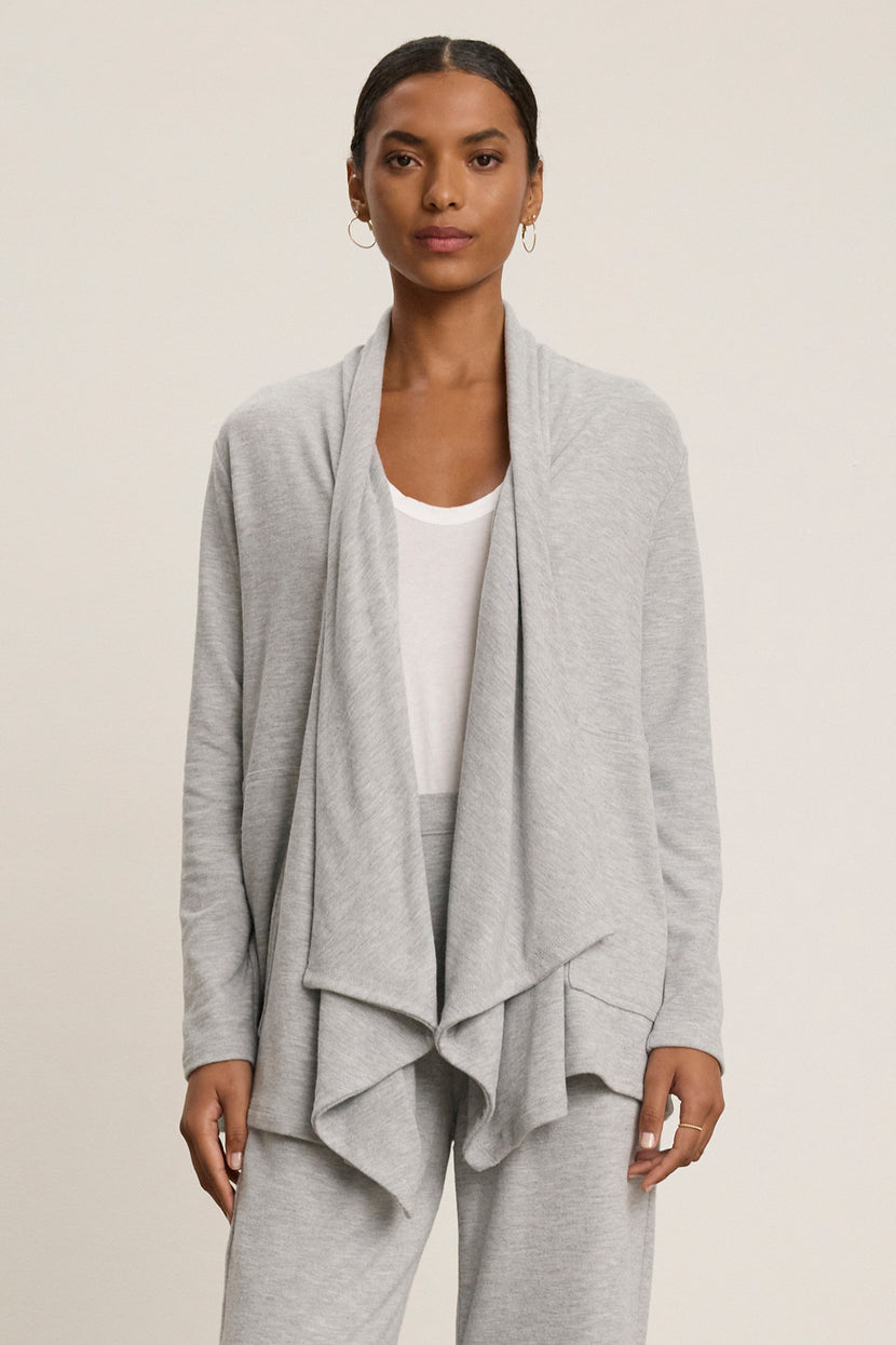 A person wearing a grey LISA CARDIGAN by Velvet by Graham & Spencer, crafted from luxurious triblend knit fabric with side patch pockets, is styled over a white shirt and matching grey pants, standing against a neutral background.