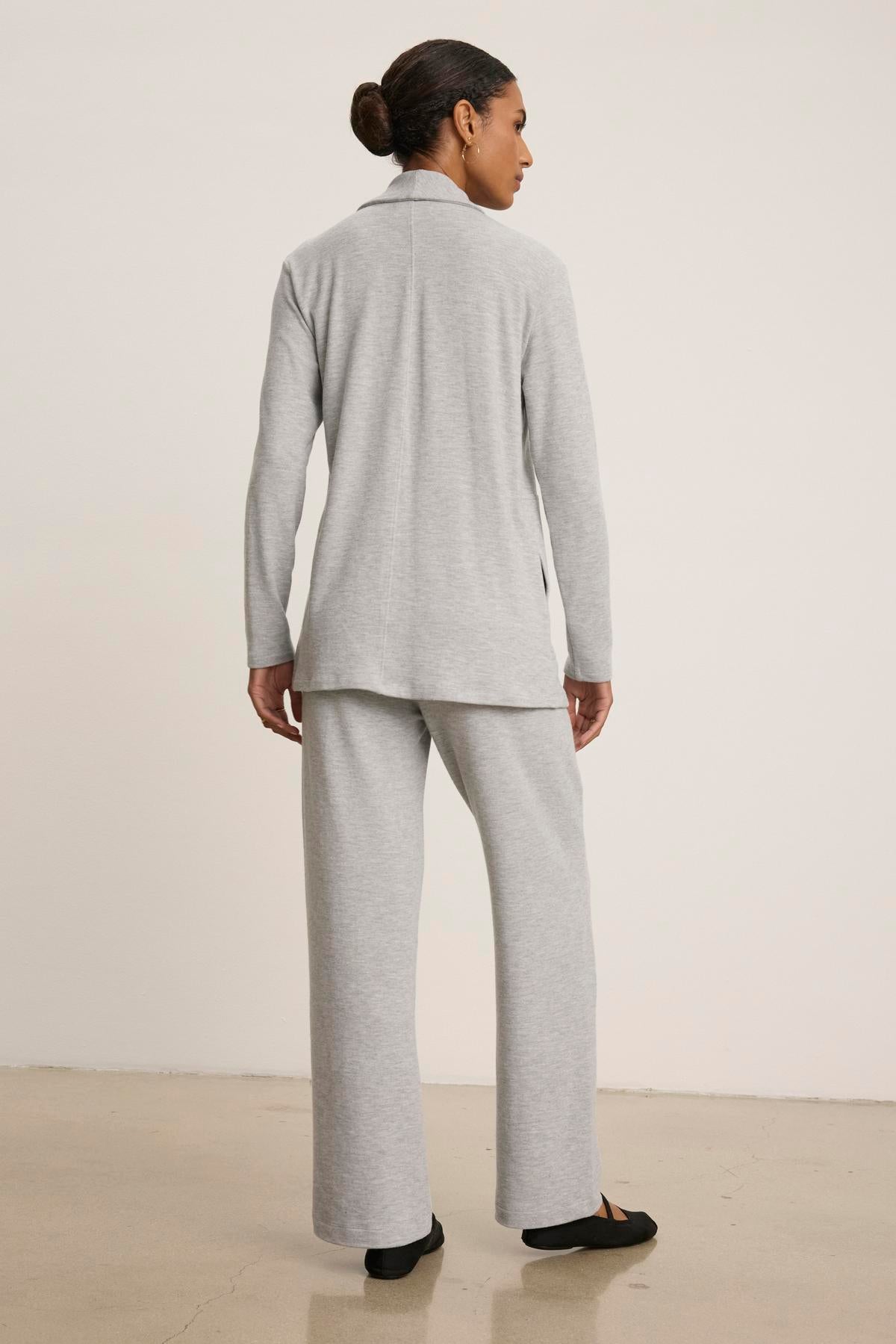   A person stands facing away, wearing the Velvet by Graham & Spencer LISA CARDIGAN pantsuit in a light gray luxurious triblend knit, featuring an open front top and wide-leg pants. Their hair is tied back, and they are on a neutral floor against a white background. 