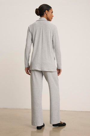 A person stands facing away, wearing the Velvet by Graham & Spencer LISA CARDIGAN pantsuit in a light gray luxurious triblend knit, featuring an open front top and wide-leg pants. Their hair is tied back, and they are on a neutral floor against a white background.