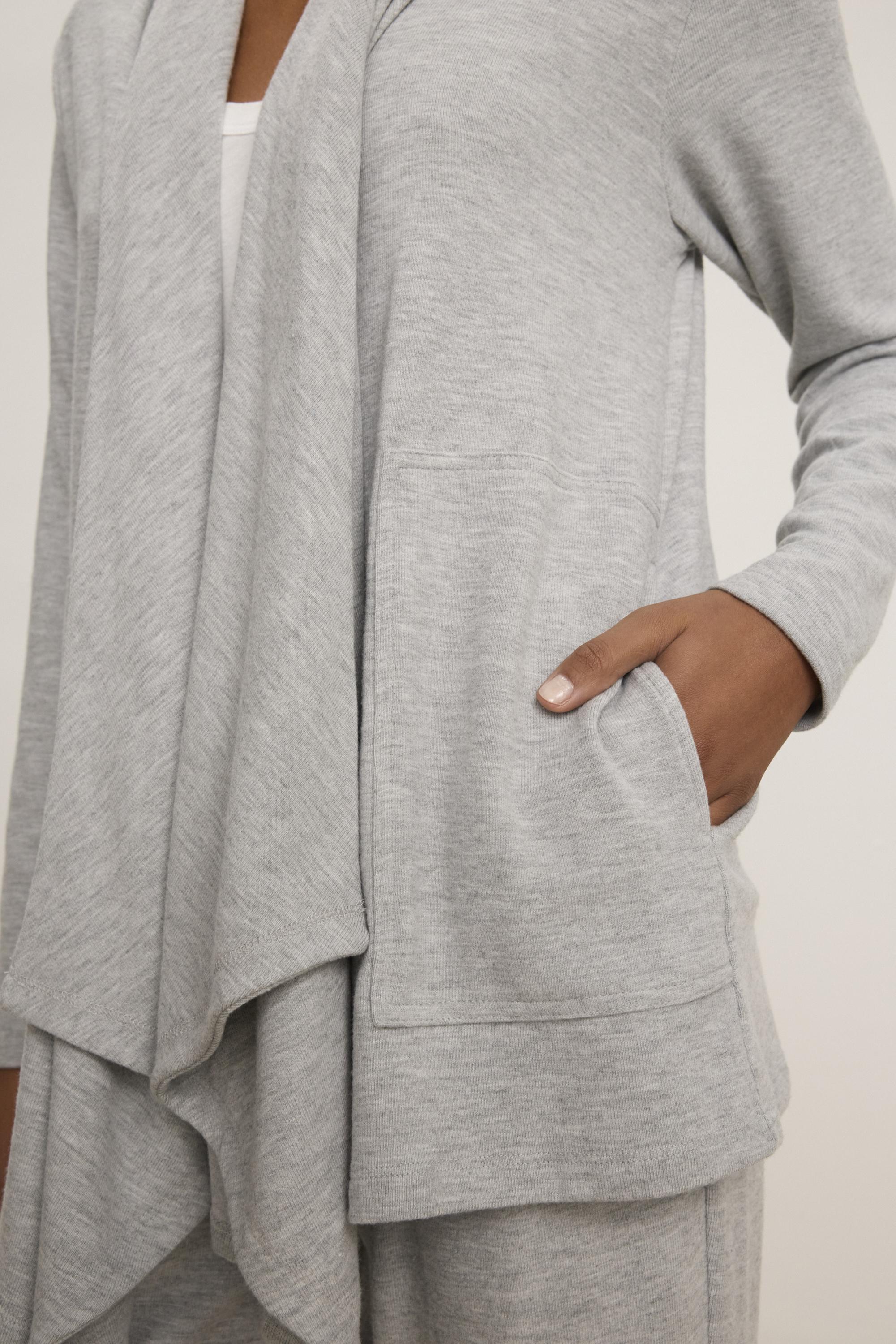   A close-up shows a person wearing the LISA CARDIGAN by Velvet by Graham & Spencer, featuring a light gray, draped design with side patch pockets in a luxurious triblend knit, and styled with one hand in the pocket over a white top. 