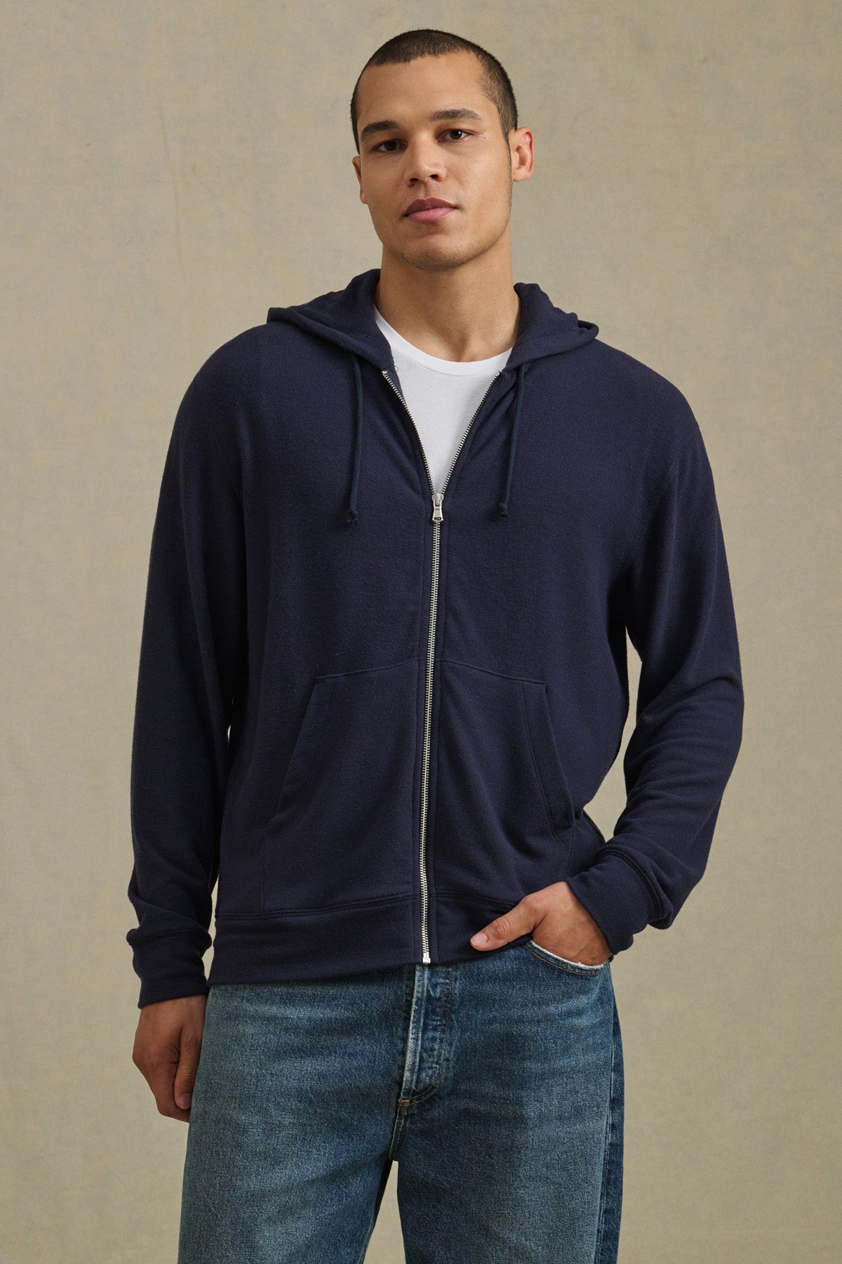 A person models the PORTER HOODIE by Velvet by Graham & Spencer in navy, layered over a white tee and blue jeans against a beige backdrop, highlighting an effortless casual style.-41009056776385