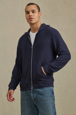 A person faces forward in the Velvet by Graham & Spencer PORTER HOODIE—navy blue, full-zip—layered over a white tee and blue jeans with one hand in the pocket, showcasing effortless, casual style.