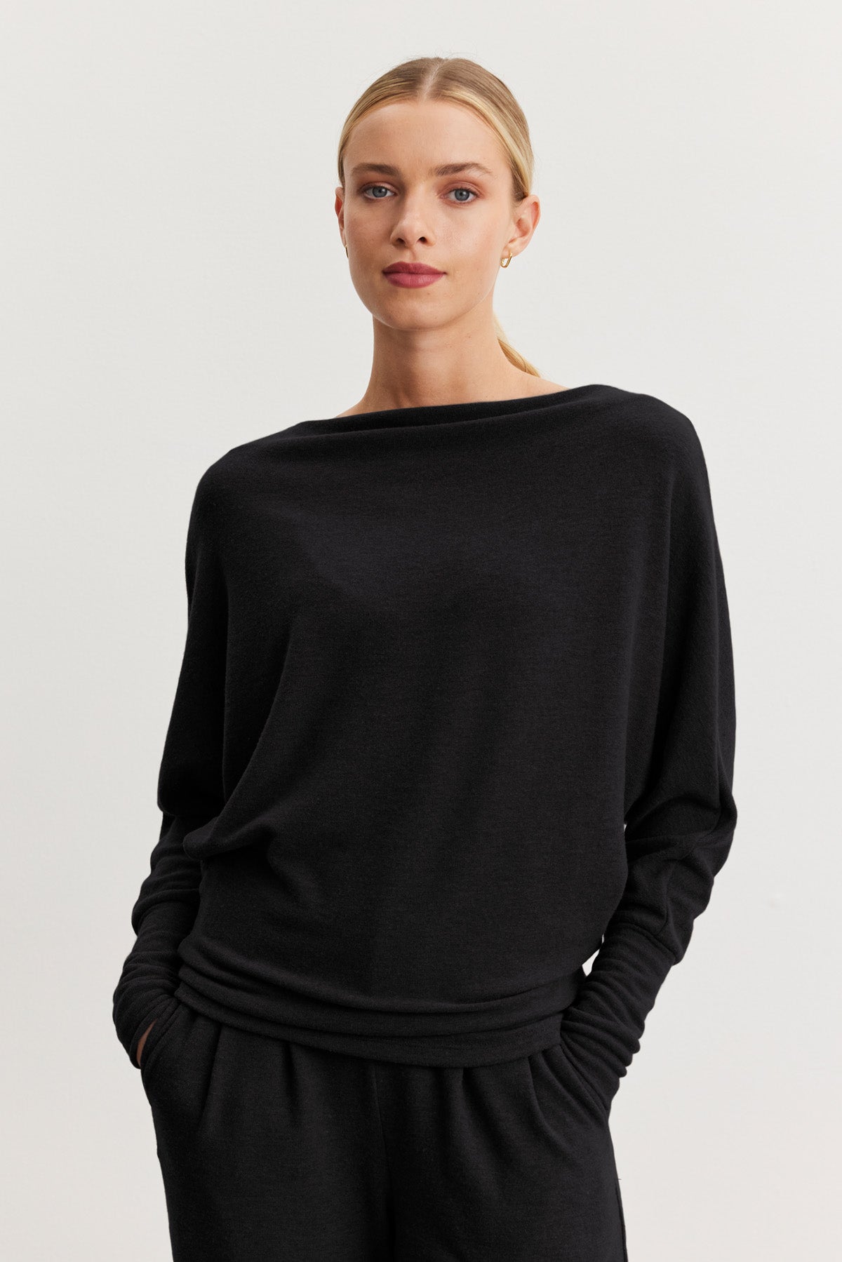 A woman with blonde hair in a low bun wears the SELMY TOP by Velvet by Graham & Spencer, a loose-fitting black triblend knit with dolman sleeves, paired with black pants against a plain light background.-40461709148353