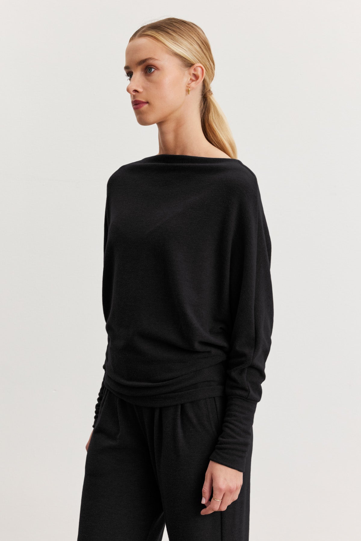 A woman with blonde hair in a low ponytail models the SELMY TOP by Velvet by Graham & Spencer, a loose-fitting black triblend knit with dolman sleeves, paired with matching black pants against a plain white background.-40461709115585
