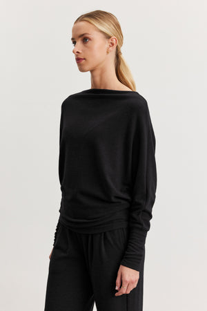 A woman with blonde hair in a low ponytail models the SELMY TOP by Velvet by Graham & Spencer, a loose-fitting black triblend knit with dolman sleeves, paired with matching black pants against a plain white background.