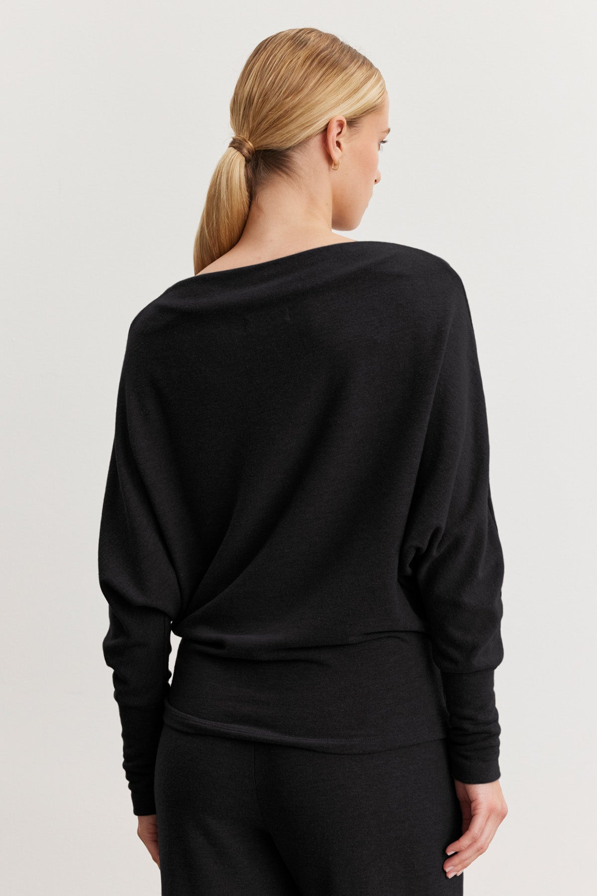 A woman with blonde hair in a low ponytail is seen from behind wearing the SELMY TOP by Velvet by Graham & Spencer, a loose-fitting black triblend knit long-sleeve top with dolman sleeves and matching black pants.-40461709082817