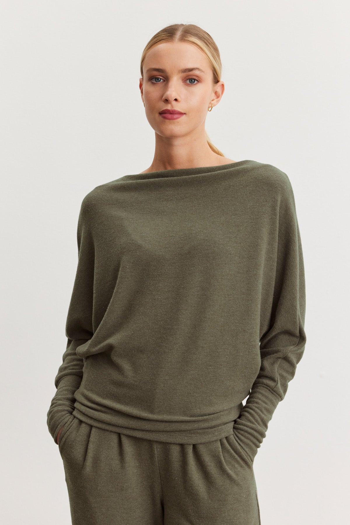 A woman with blonde hair tied back wears the SELMY TOP by Velvet by Graham & Spencer in olive green, paired with matching pants, standing against a plain light background with her hands in her pockets.
