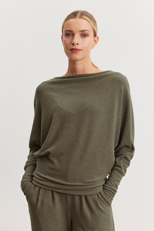 A woman with blonde hair tied back wears the SELMY TOP by Velvet by Graham & Spencer in olive green, paired with matching pants, standing against a plain light background with her hands in her pockets.