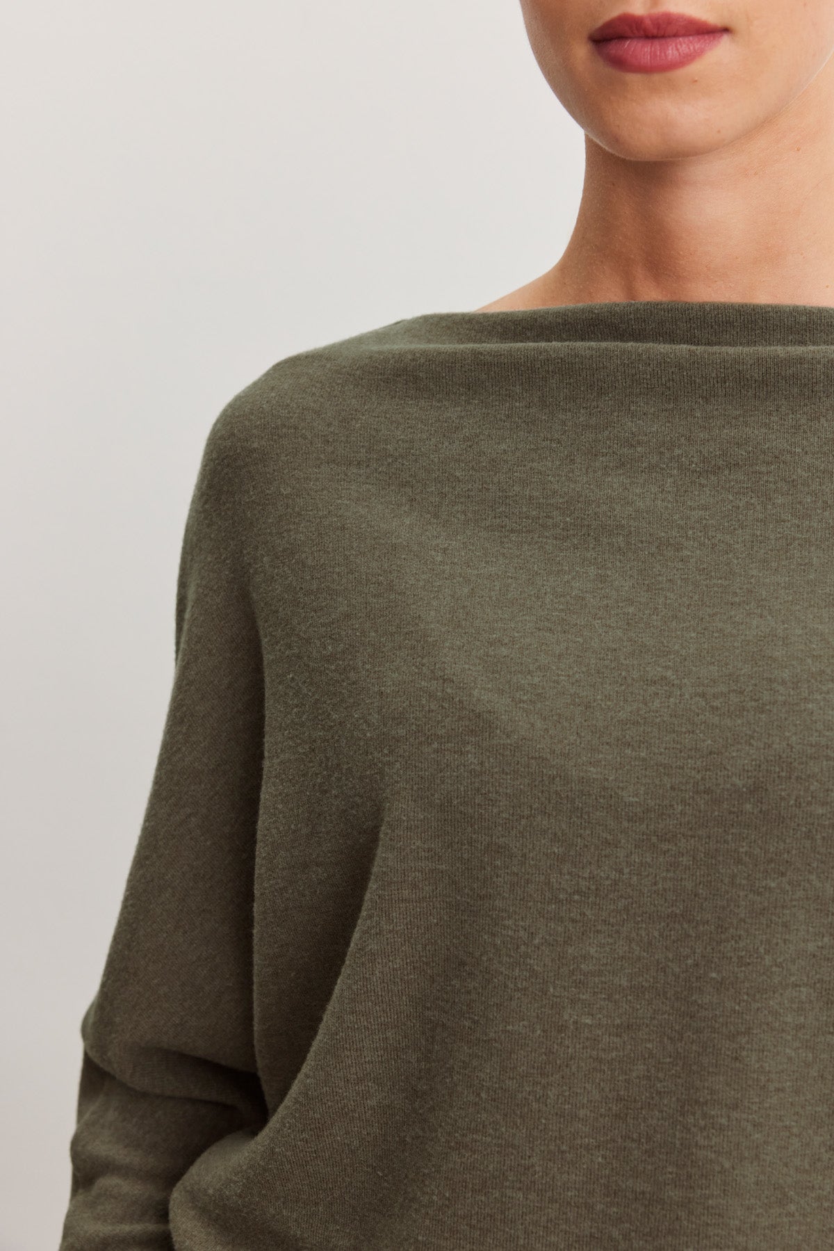 A close-up shows a person wearing the SELMY TOP by Velvet by Graham & Spencer, an olive green triblend knit boat neck with long dolman sleeves; only the lower face and upper torso are visible.-40500009238721