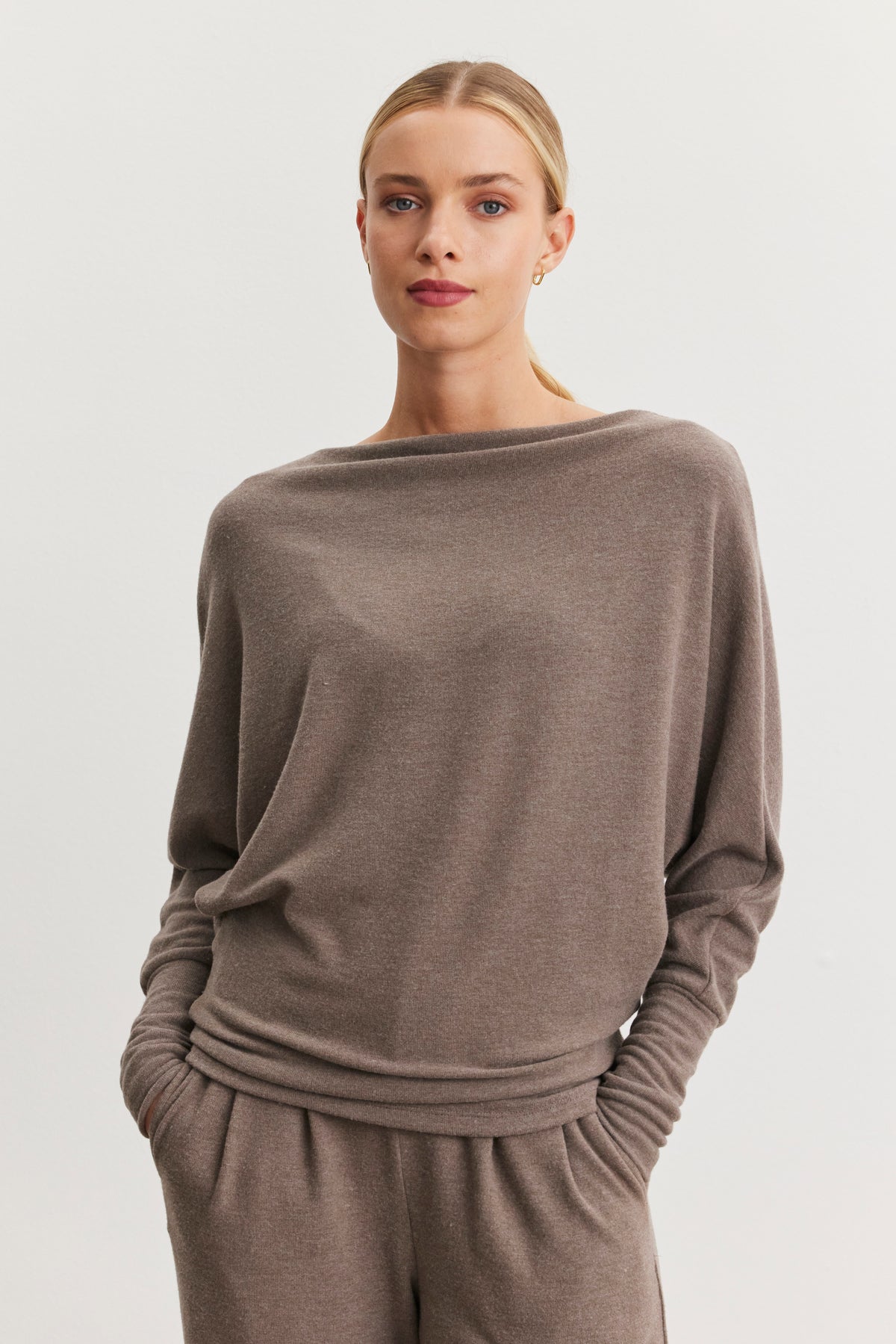   The individual is posed against a simple, light backdrop, with hands tucked into the pockets of a loose, taupe sweater called the SELMY TOP by Velvet by Graham & Spencer. This piece features a boat neck and dolman sleeves, with its soft triblend knit enhancing the comfort of the relaxed ensemble. 