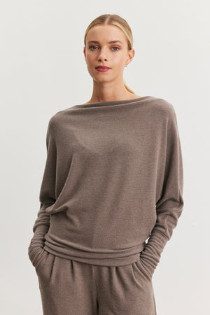 The individual is posed against a simple, light backdrop, with hands tucked into the pockets of a loose, taupe sweater called the SELMY TOP by Velvet by Graham & Spencer. This piece features a boat neck and dolman sleeves, with its soft triblend knit enhancing the comfort of the relaxed ensemble.