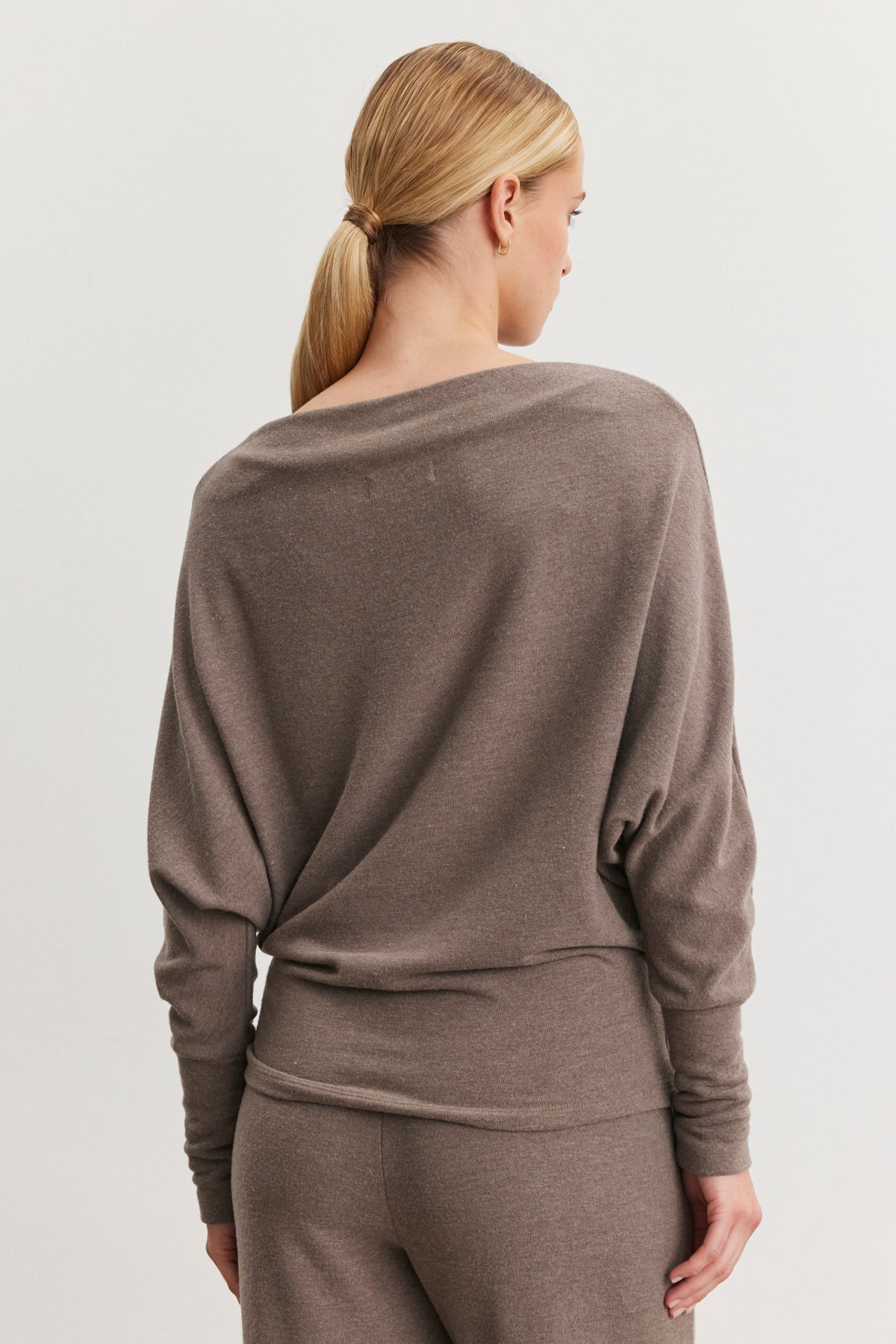   A person with blonde hair in a low ponytail is facing away, wearing the Velvet by Graham & Spencer SELMY TOP in taupe, featuring dolman sleeves and matching pants. The soft triblend knit enhances her outfit's understated elegance. 