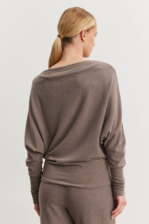 A person with blonde hair in a low ponytail is facing away, wearing the Velvet by Graham & Spencer SELMY TOP in taupe, featuring dolman sleeves and matching pants. The soft triblend knit enhances her outfit's understated elegance.