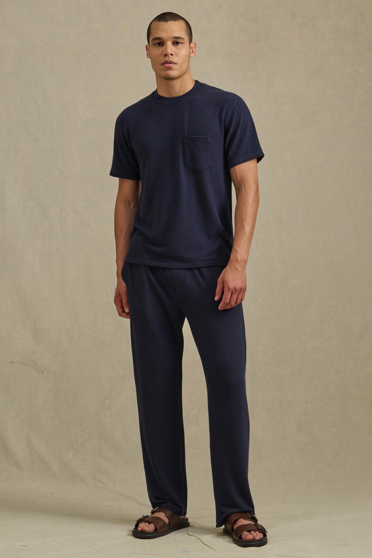 A man stands against a beige background wearing a navy tri-blend short-sleeve T-shirt with a chest pocket, EASTON SWEATPANT by Velvet by Graham & Spencer in matching navy, and brown sandals.