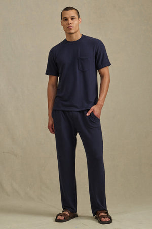 A person stands against a plain background wearing the Velvet by Graham & Spencer EASTON SWEATPANT in navy with an elastic waist, paired with a navy short-sleeve shirt featuring a chest pocket and brown sandals.