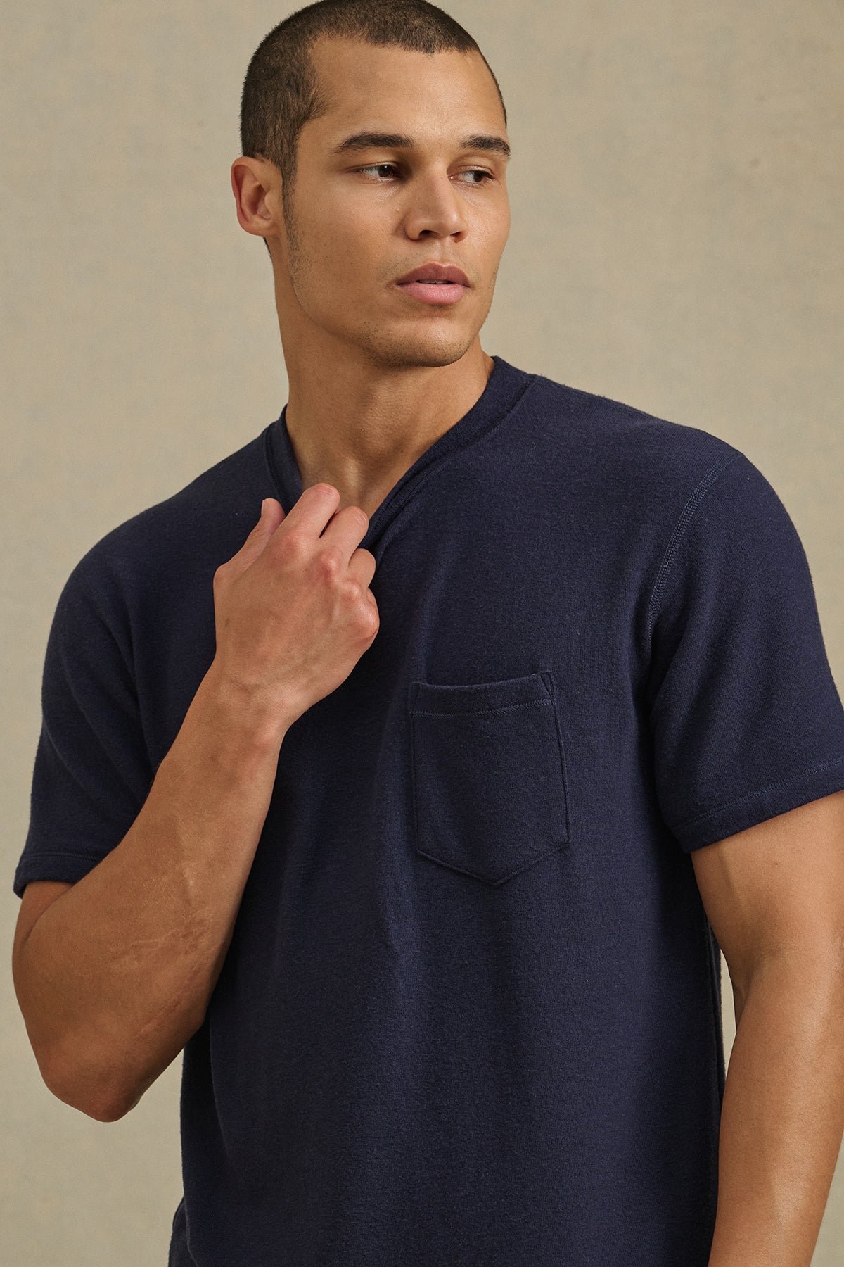 A man wears the SIMON CREW short-sleeve shirt by Velvet by Graham & Spencer in navy blue tri-blend fabric with a chest pocket, holding the collar and looking to his left against a plain background.-41008422125761
