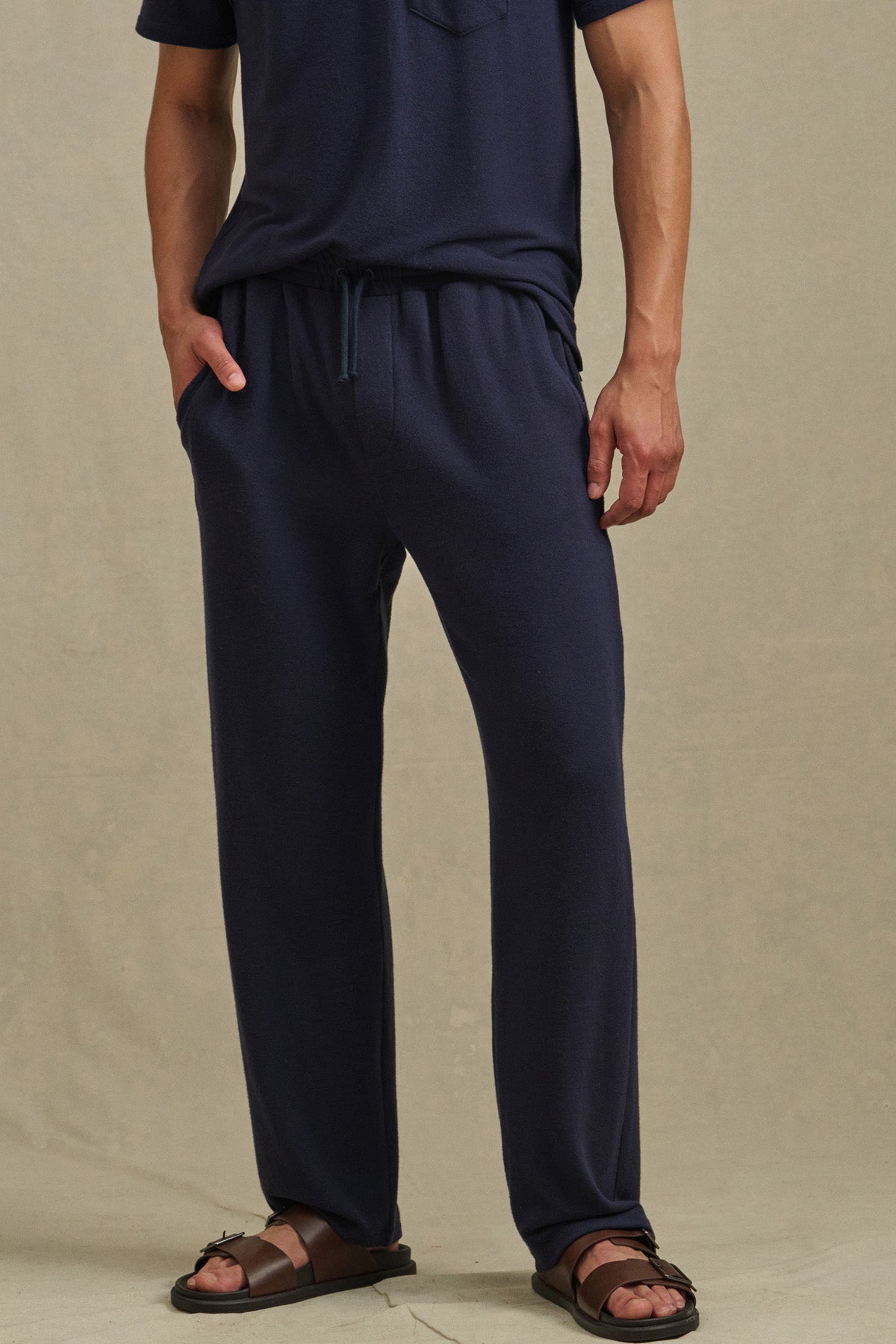 A person wearing Velvet by Graham & Spencer's EASTON SWEATPANT and a navy blue t-shirt with brown sandals stands against a plain beige background.
