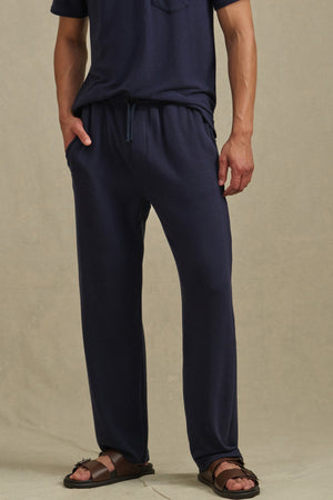 A person wearing Velvet by Graham & Spencer's EASTON SWEATPANT and a navy blue t-shirt with brown sandals stands against a plain beige background.