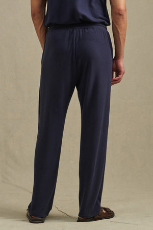 A person stands with their back to the camera, wearing Velvet by Graham & Spencer’s EASTON SWEATPANT in navy, paired with a matching shirt and brown sandals.