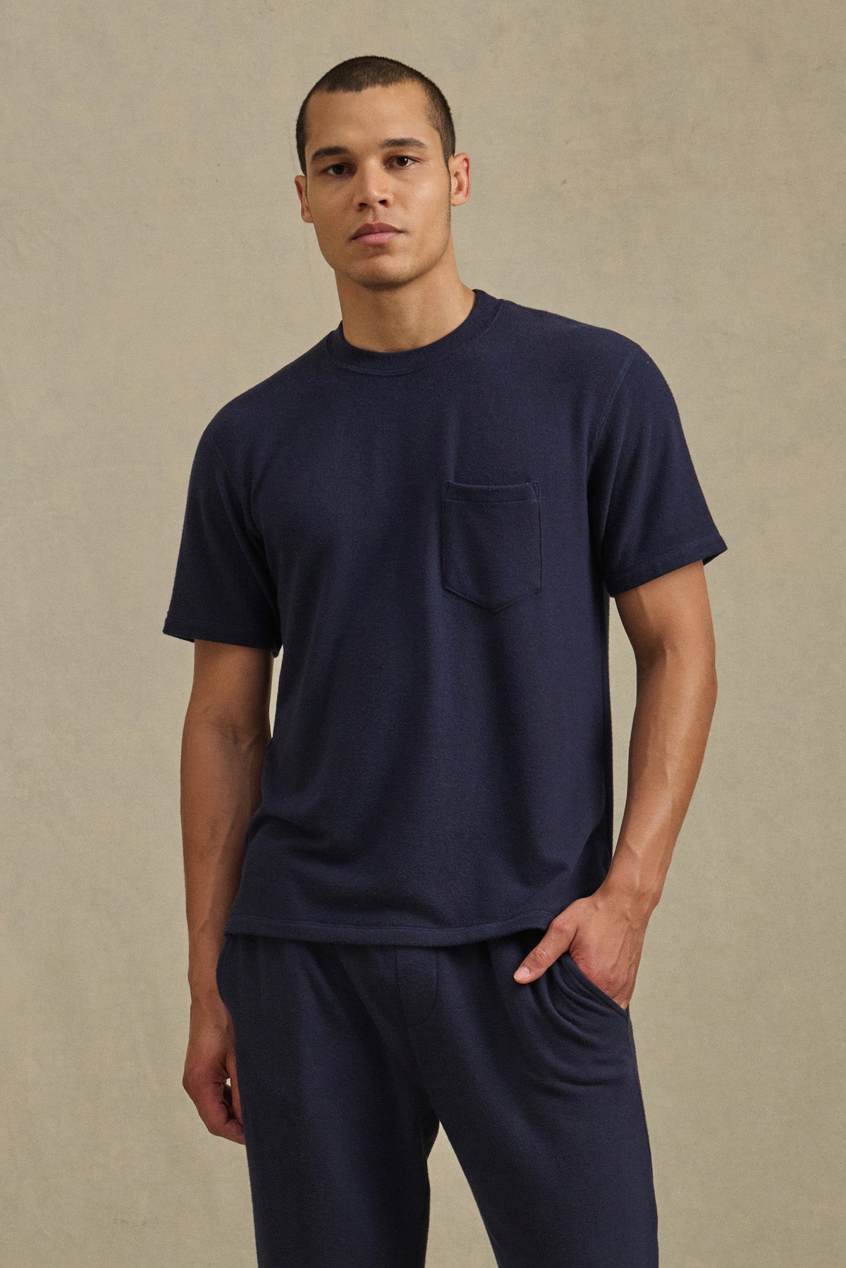 A man wears the SIMON CREW short-sleeve navy t-shirt with chest pocket by Velvet by Graham & Spencer, paired with matching navy pants, standing against a neutral background.-41009058939073