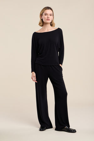 A person stands against a beige background, looking at the camera with a neutral expression, wearing the Velvet by Graham & Spencer MACKENZIE LOUNGE PANT and a matching loose-fitting black long-sleeve top crafted from soft knit fabric, paired with black shoes.