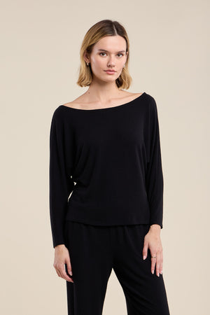 A woman wears the Velvet by Graham & Spencer CECILIA TOP—a black, long-sleeve, boat neck style crafted from soft knit fabric—paired with matching relaxed-fit black pants, standing against a plain beige background.