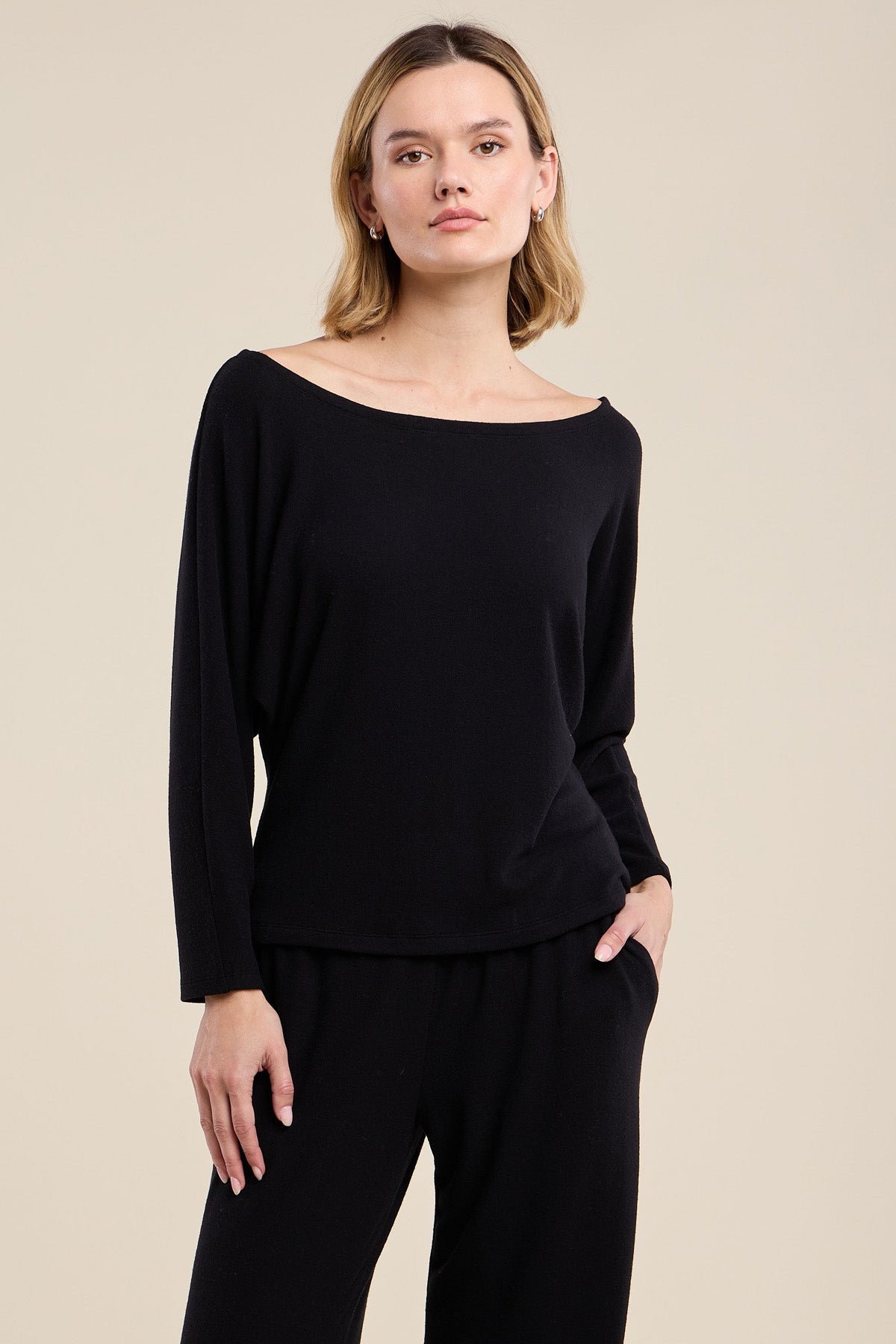 A person wearing the CECILIA TOP by Velvet by Graham & Spencer— a black, relaxed fit long-sleeve top —and black pants stands against a plain beige background, facing forward with a neutral expression.
